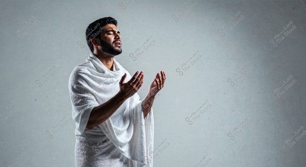 An image of a man wearing traditional white Ihram clothing, appearing contemplative and serene, with his hands raised in prayer. The background is a simple gray, highlighting the figure.