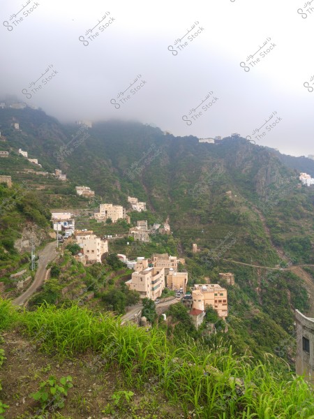 A landscape view of houses built on steep, green-covered mountain slopes. Low clouds partially cover the mountains, giving the scene a foggy appearance. Narrow roads wind between the houses, with lush vegetation filling the foreground.
