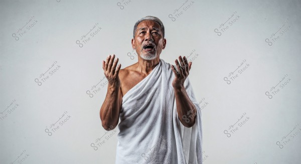 An elderly man with white hair and beard standing against a white background. The man is wearing an Ihram, a traditional garment worn during the Islamic pilgrimage of Hajj or Umrah. He is in a posture with raised hands, likely in prayer.