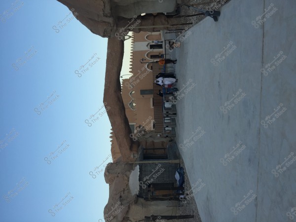 The image shows an archaeological site with large stone structures featuring traditional design and clay buildings in the background. Several people are present, wearing traditional clothing, likely in a Middle Eastern setting.