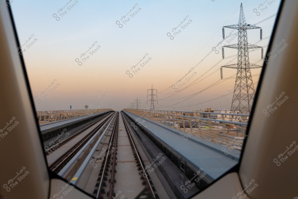 A view through a train window showing railway tracks extending towards the horizon during sunset. There are electricity pylons on the right side, and the sky is a blend of light blue and orange colors. The scene conveys a sense of calm and technological advancement.