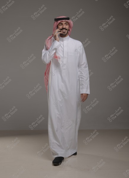 A portrait of a man wearing a white thobe, a red and white checkered ghutra, and a black agal, standing and smiling while talking on the phone. The attire suggests traditional Saudi clothing, and the background is a neutral gray color.