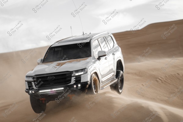 A white SUV driving quickly across sand dunes in the desert, kicking up dust around it. The sky is partially cloudy in the background.