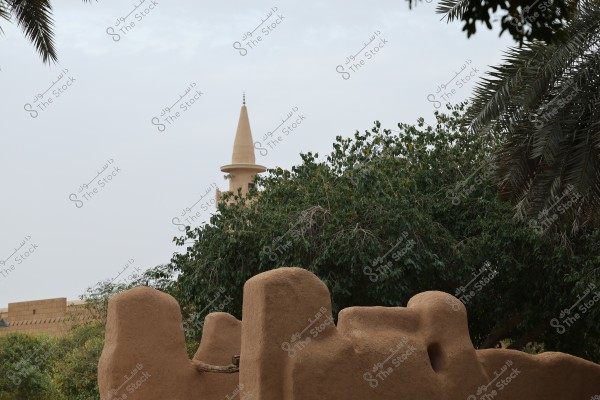 The image shows a historical mud structure with a part of a minaret or castle peeking in the background. Dense trees and palm trees surround the structure, indicating a natural setting. The sky is cloudy, adding a serene atmosphere to the scene.