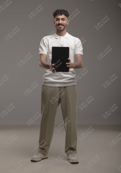 An image of a man standing in a studio. He is wearing a white polo shirt, beige pants, and sneakers. He holds a black tablet with both hands and smiles while looking at the camera. The background is gray and neutral.
