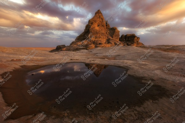 The image shows a rocky mountain in the desert under a cloudy sky during sunset. The mountain is faintly reflected in a small puddle of water on the flat rocky ground in front of it. The setting sun casts warm colors on the mountain peak, while the horizon appears distant and open.