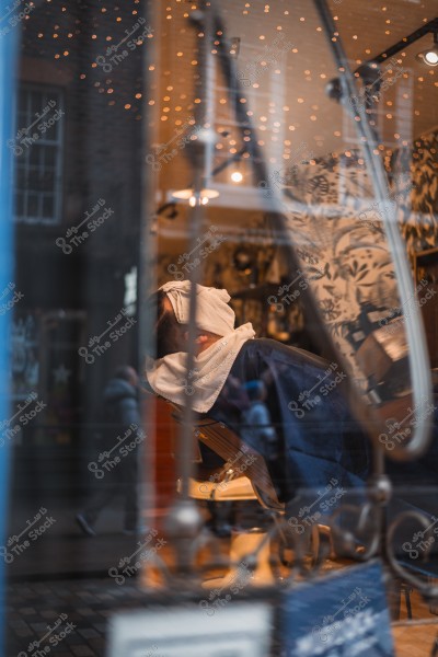 Image of a man relaxing in a barbershop, with a towel on his face, and lights reflecting on the shop window from outside.