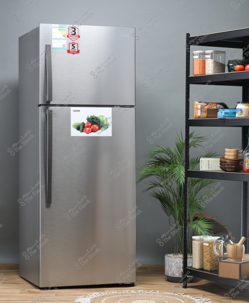 An image of a modern kitchen featuring a large silver refrigerator with a sleek and stylish design, with a sticker on the door displaying vegetables like tomatoes and lettuce. Next to the refrigerator are storage shelves holding transparent containers with lentils and grains, along with wooden bowls. Indoor plants in pots are placed on the floor, adding a natural touch to the interior decor.