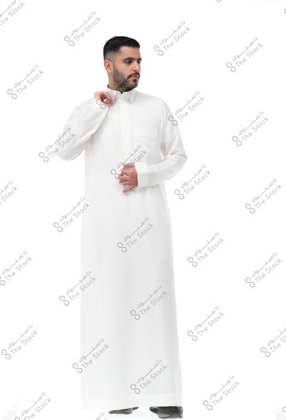 Image of a man wearing a long white thobe with long sleeves and a collar, which is traditional clothing in the Gulf region. The man stands against a white background and is holding his collar with his right hand.