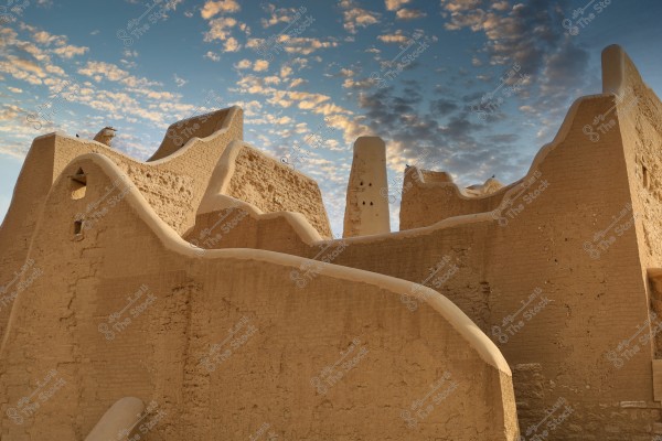 View of a traditional mud-brick building in Saudi Arabia, showcasing the light brown walls built from clay bricks. The walls feature sculpted designs with prominent architectural details. The sky behind the building is blue with scattered clouds, which adds an aesthetic touch to the scene.