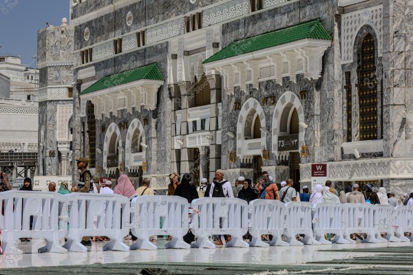 The image shows a group of people standing in front of one of the entrances to the Grand Mosque in Mecca. The individuals are dressed in various traditional Islamic attire. A white plastic barrier separates the crowd from the building. The architecture features intricate Islamic designs with arches. The sky above is clear.