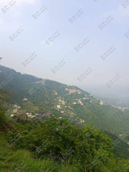A natural landscape of green terraced mountains covered with vegetation and a scattering of houses on the mountain slopes. The sky appears partially cloudy, with clouds covering the distant mountain tops, giving a hazy appearance to the horizon.