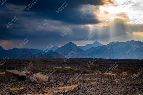 A natural landscape showing a mountain and rocky terrain under a cloudy sky. The sun peeks through the clouds, casting rays of light on the mountains in the background. The ground is covered with rocks and scattered vegetation.
