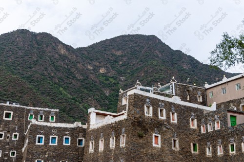 An ancient building built of stones with a group of windows surrounded by white paint in the middle, a group of mountains covered with many trees while the sky looks cloudy during the day, Rijal Almaa heritage village in the Asir region