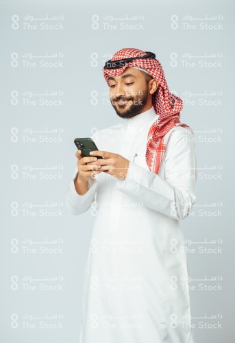 بورتريه رجل سعودي مبتسم يرتدي الزي السعودي التقليدي يستخدم الجوال وينظر للشاشة بينما يتصفحه