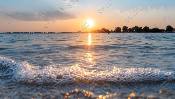 A sunset view over the sea with orange sunlight reflecting on the calm water and small waves gently lapping on the sandy shore. Trees are visible on the horizon under a sky with light clouds.