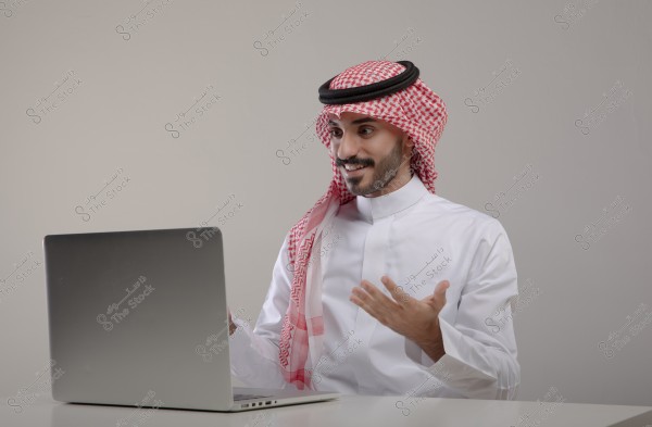 The image shows a man wearing a white thobe, red keffiyeh, and agal, sitting in front of a laptop. He appears to be in a discussion or video call, with expressive gestures and expressions on his face.