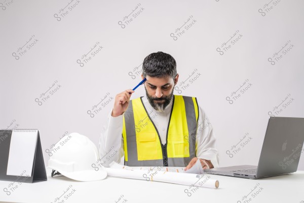 Image of a man wearing a yellow reflective vest, working at a desk with a white helmet, a calendar notebook, and a laptop. He appears to be reviewing blueprints, holding a blue pen to his head in deep thought. The man is wearing traditional white clothing and seems to be in an office work environment.