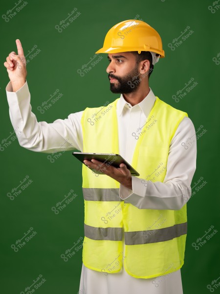 A photo of a man wearing a yellow construction helmet and a yellow reflective safety vest, standing in front of a green background. He is dressed in a traditional white Middle Eastern garment and holds a tablet in his left hand while pointing upwards with his right finger. The portrait suggests Middle Eastern origin.