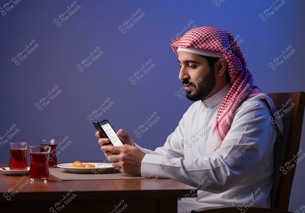 ` A man sitting at a table wearing a traditional white thobe and a red and white shemagh. He is holding a smartphone and looking at it attentively. In front of him on the table is a plate with some pastries and two glasses of tea. The background is deep blue, highlighting the details of the scene.\r\n\r\n`