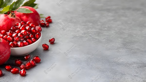 An image of four ripe, red pomegranates placed side by side on a gray background. A portion of the shiny, jewel-like seeds is displayed in a small white bowl near the pomegranates, with some seeds scattered around it.