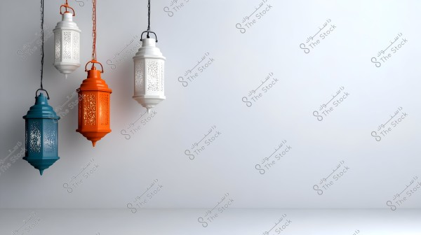 A collection of traditional Arabic lanterns hanging on a wall. The lanterns are in various colors including white, orange, and dark blue, featuring decorative engravings that are highlighted when lit. The background is a plain, light-colored wall.