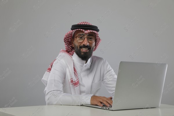 صورة لرجل يرتدي ثوباً أبيض تقليدياً وغترة حمراء وبيضاء مع عقال, يجلس أمام حاسوب محمول على مكتب. يبدو سعيداً ومرتاحاً وخلفيته رمادية بسيطة.