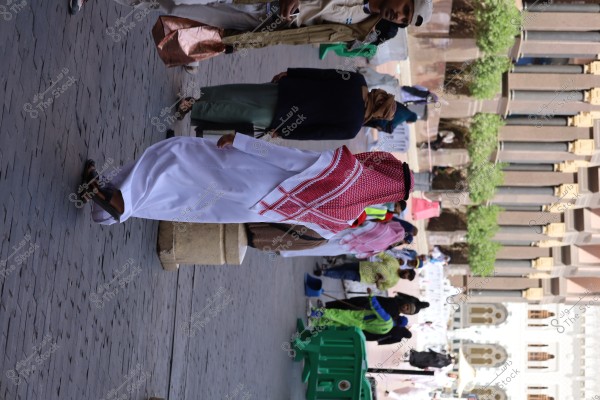 A man wearing a white thobe and a red keffiyeh walking in a public area, possibly in Saudi Arabia, with other people in the background. A building with traditional architectural design is visible on the side.\r\n\r\n###
