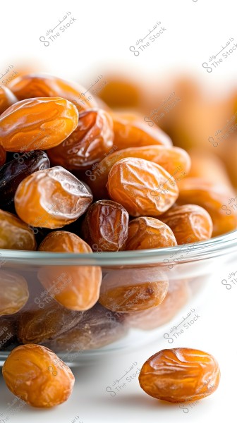 An image showing a collection of brown dates in a clear glass bowl. The dates are glossy and deep brown, of medium size, showcasing the crinkled texture on their surface. Some dates are interlaced in a way that shows the color variations between light and dark brown against a blurred white background.