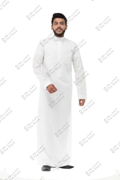 A portrait of a man standing, wearing a long-sleeved white thobe, possibly of Saudi origin, with a smiling expression. He is wearing black shoes against a simple white background.