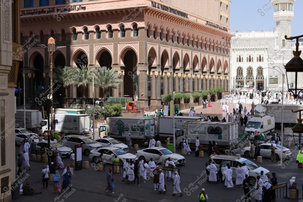 An image of a large architectural complex overlooking a busy square in Medina, Saudi Arabia. The image shows several people wearing traditional white Ihram clothing, along with cars and trucks with signs written in Arabic. The surrounding buildings feature arches and numerous windows. Palm trees are visible in the open area, and many people are gathering in the large square next to the buildings. In the background, part of the Prophet\'s Mosque is visible.