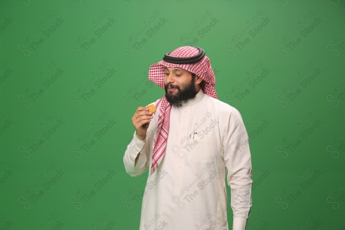 شاب سعودي على خلفية خضراء يرتدي الثوب و الشماغ، يقوم بعمل حركات بيده،يمسك الكعك و يأكله ، احتفالات العيد