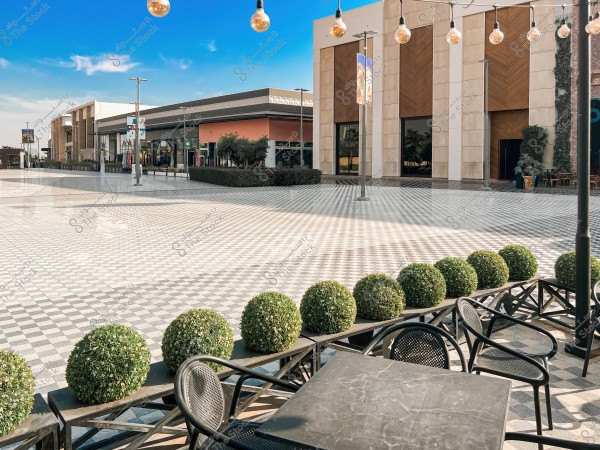 A wide outdoor plaza with tables and chairs in the foreground. A row of round shrubs lines a metal barrier. In the background, modern commercial buildings with large glass windows are visible. Small lights are hung across the plaza, and the sky is clear blue.