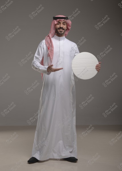 Image of a man wearing a white thobe and a red checked ghutra with an agal. He stands smiling in a welcoming pose, holding a white circular board. The background is plain gray.