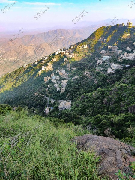 A scenic view of a mountain village in the Asir region of Saudi Arabia. The image features scattered houses built on green terraces along the mountain slope. In the background, expansive mountain ranges extend under a clear blue sky.