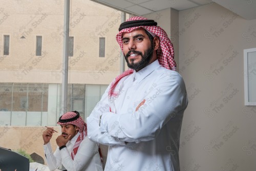 رجل سعودي يرتدي الزي السعودي التقليدي يقف مكتوف اليدين أثناء إجتماع في العمل نهارًا