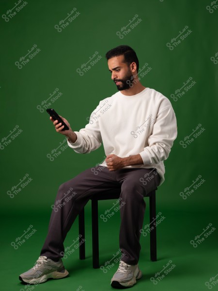 A photo of a man sitting on a chair against a green background. He is wearing a white sweater, gray sweatpants, and gray sneakers. He is looking at his mobile phone in his right hand with a focused expression.