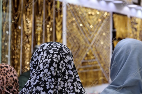 The image shows two women wearing headscarves; one wears a headscarf with black and white floral patterns, and the other in light blue. Behind them are golden wall hangings and decorations, resembling a gold or jewelry display.