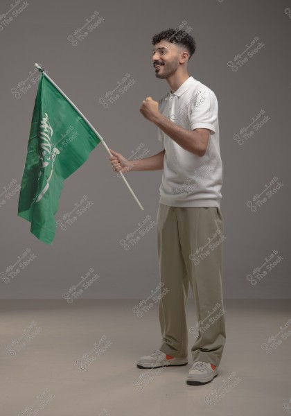 Image of a man standing and waving the flag of Saudi Arabia. He is wearing a white shirt, beige pants, and sneakers, appearing happy and enthusiastic. The background is grey and neutral, highlighting the man\'s posture and the green Saudi flag.