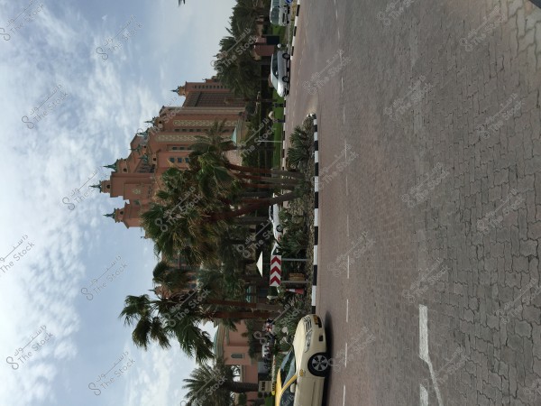 Image of a large building extending into the background with ornate towers and a pink facade, surrounded by a variety of trees and palm trees. In the foreground, part of a roundabout is visible on the road with a white car and a yellow car parked nearby. The sky is dotted with light clouds.