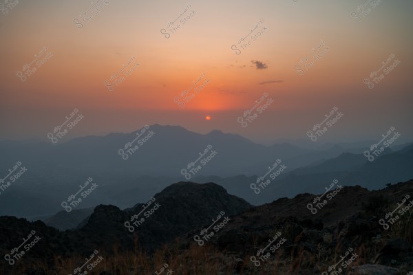 A view of a sunset over misty mountains. The horizon displays a gradient of orange and yellow, while the mountains in the foreground appear dark and their features indistinct.