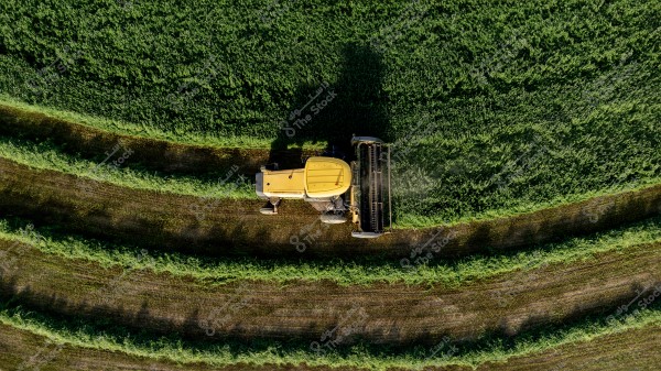 Aerial view of a yellow agricultural harvester moving through a green, planted field. The machine is cutting the grass in parallel rows, leaving a clear pattern in the field.