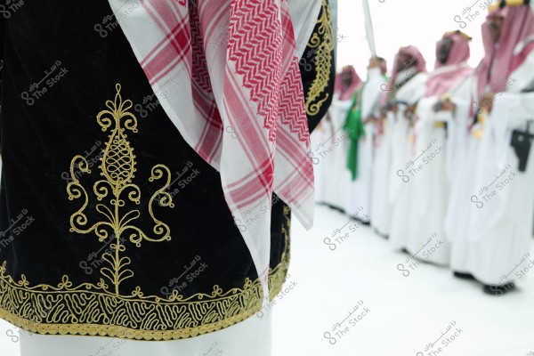 The image shows a group of people wearing traditional clothing, focusing on a black garment embroidered with a golden design, and a red and white checked scarf typically associated with Saudi Arabian attire. In the background, men are standing wearing white robes and red-checked scarves.