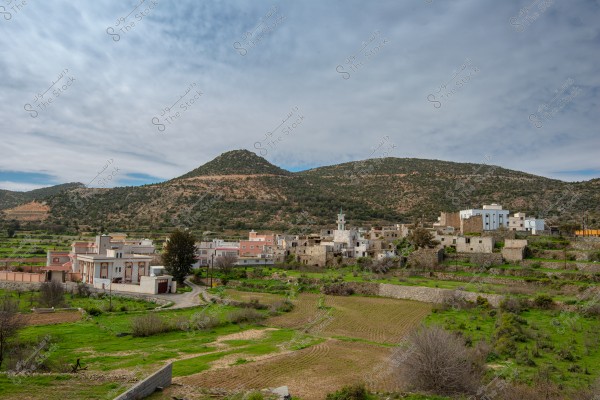 صورة لمنطقة ريفية تُظهر مجموعة من البيوت والمباني التقليدية المتراصة صفًا في قرية, محاطة بمساحات خضراء وحقول زراعية. تظهر في الخلفية تلال مغطاة بالنباتات, وتحت السماء الزرقاء المغطاة بالغيوم الخفيفة. يظهر مسجد صغير في منتصف القرية.