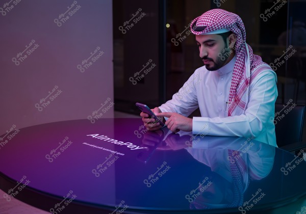 Image of a man seated at a round table with a sleek appearance, wearing traditional thobe and ghutra. The man is using a mobile phone, with the name \"AlimnaPay\" displayed on the table surface. The scene is illuminated with blue and purple lights, creating a modern atmosphere.