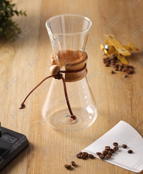 A glass and wood coffee brewing carafe on a wooden table, with ground coffee, a filter paper, and a small toy airplane beside it.