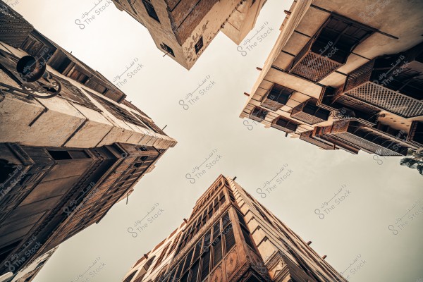 صورة تظهر منظرًا لأربعة مبانٍ مائلة إلى السماء بتصميم تقليدي يُظهر النوافذ والشرفات البارزة. الجدران تبدو أثرية وتدل على طراز معماري قديم معقد, يمكن أن يكون من منطقة عربية مثل جدة, السعودية. السماء صافية مع ضوء دافئ يعكس تفاصيل المباني.