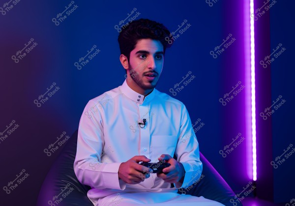 A portrait of a man sitting on a beanbag chair in a dimly lit room with purple and blue neon lights. The man is wearing a white thobe, suggesting Saudi origin, and is holding a game controller. His expression shows focus and engagement in his activity.