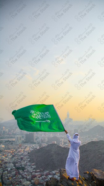 A snapshot of a Saudi man in traditional Saudi dress holding the Saudi flag over a mountain in the Makkah Al-Mukarramah region, the sky, the royal clock tower building in the Grand Mosque, buildings and landmarks, the Grand Mosque of Mecca.