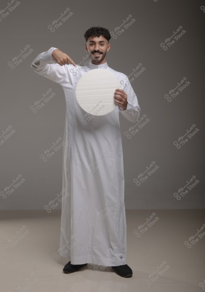Image of a man wearing a white thobe pointing at a white circle he holds in his hand. He stands against a gray backdrop and appears to be smiling. The traditional attire suggests a Gulf region origin.